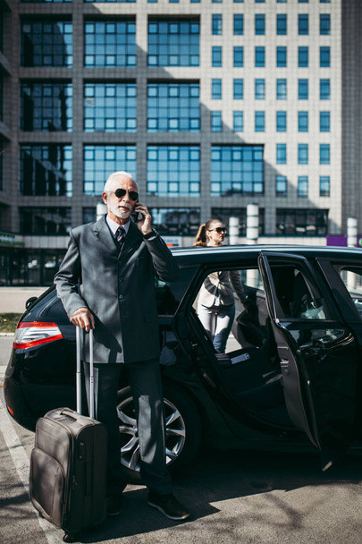 Good looking senior business man and his young woman colleague or coworker standing by the  car. He talking on smartphone, holds a suitcase and prepares to go on a business trip. Transportation in corporate business concept. - Zdjęcie, obraz