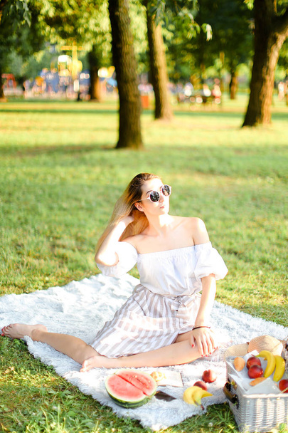 Young european girl in sunglasses having picnic on plaid and sitting in park with fruits. - Photo, image