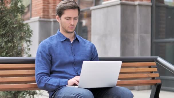 Молода людина, що працює на ноутбуці, сидячи на відкритому повітрі на лавці - Кадри, відео