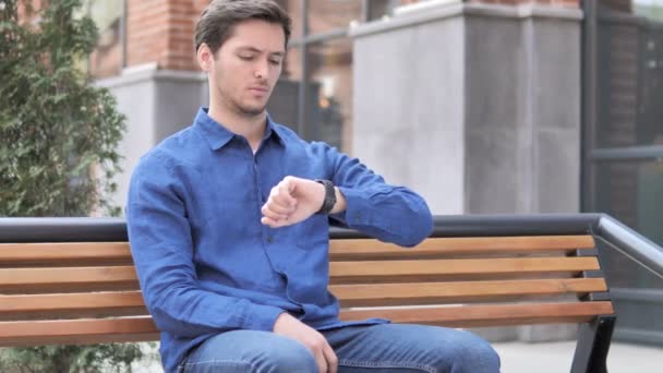 Περιμένοντας νέος άνθρωπος βλέποντας τον χρόνο στο ρολόι, καθισμένος στον πάγκο - Πλάνα, βίντεο