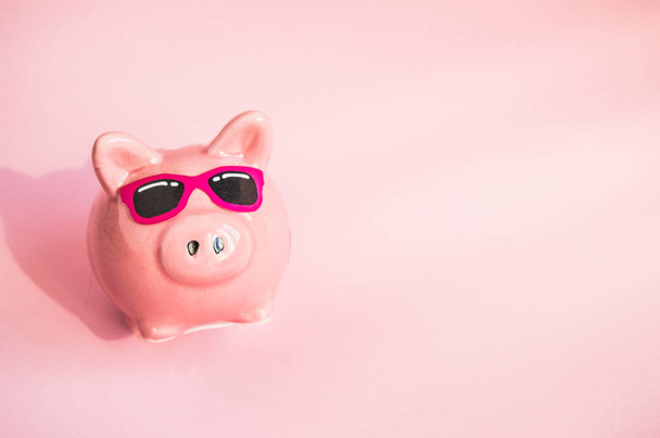 Funny Piggy Bank en gafas de sol sobre fondo rosa, luz solar, espacio para copiar, concepto de ahorro de dinero para las vacaciones de verano
 - Foto, imagen