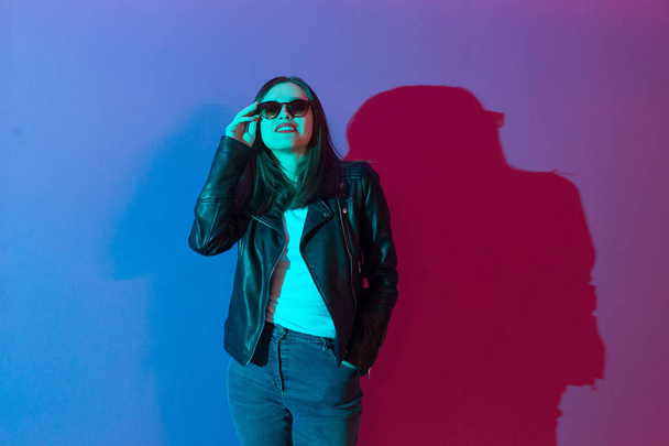 Fashioned girl in leather jacket in dark glasses in pink anf blue  neon lights background.  - 写真・画像
