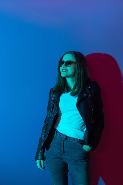 Fashioned girl in leather jacket in dark glasses in pink anf blue  neon lights background.  - Zdjęcie, obraz