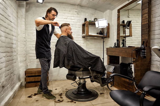 Um brutamontes na Barbearia moderna. Cabeleireiro faz penteado um homem com uma longa barba. Cabeleireiro mestre faz penteado por tesoura e pente
 - Foto, Imagem