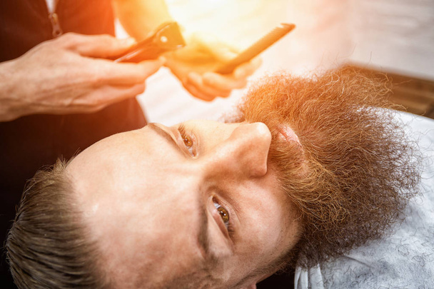 Brutalny facet w nowoczesnym salonie fryzjerskim. Fryzjer czyni fryzurę mężczyzną z długą brodą. Master fryzjer robi fryzurę nożyczkami i grzebieniem - Zdjęcie, obraz