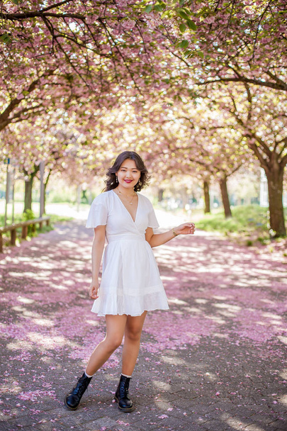Uma jovem mulher asiática bonita em um vestido branco caminha em um parque florido. Sakura. Plantas em flor
. - Foto, Imagem