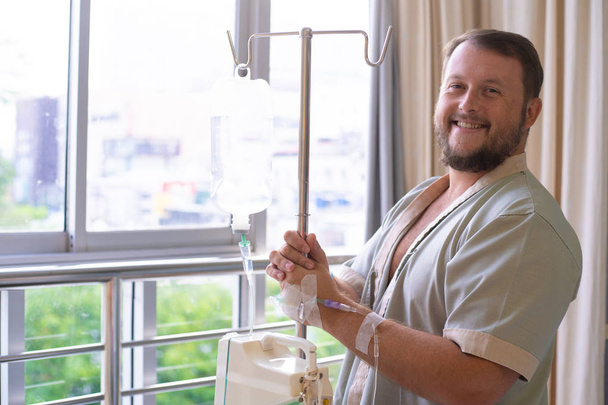 пациент с капельницей смотрит в окно больничной палаты и улыбается. Концепция здравоохранения
 - Фото, изображение