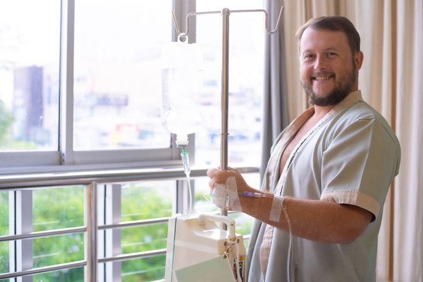 patient with a dropper looks out the window of the hospital room and smiles. Healthcare concept - 写真・画像