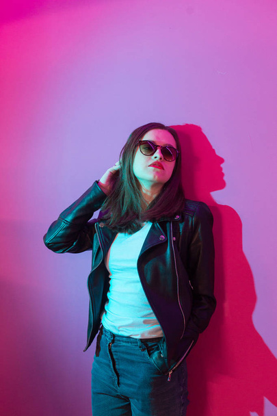 Fashioned girl in leather jacket in dark glasses in pink anf blue  neon lights background.  - Фото, изображение