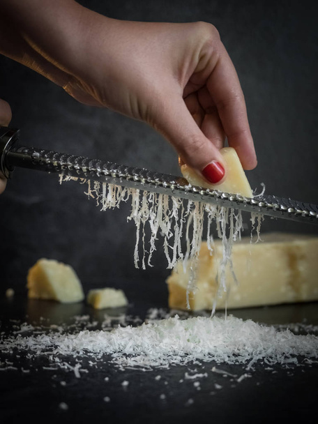 Mano de una mujer rallando queso parmesano sobre un fondo negro. Comida oscura
 - Foto, imagen