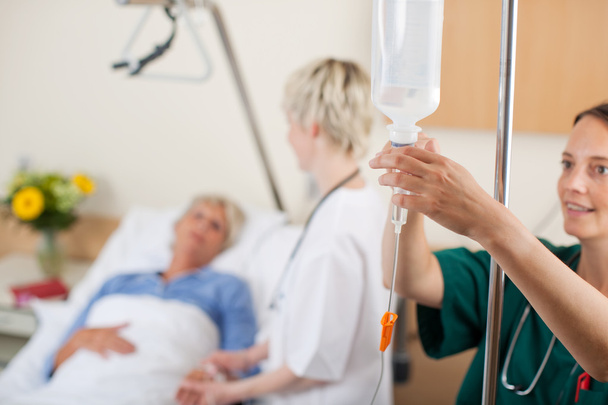 Nurse Adjusting Infusion Bottle With Doctor And Patient In Background - Foto, Bild