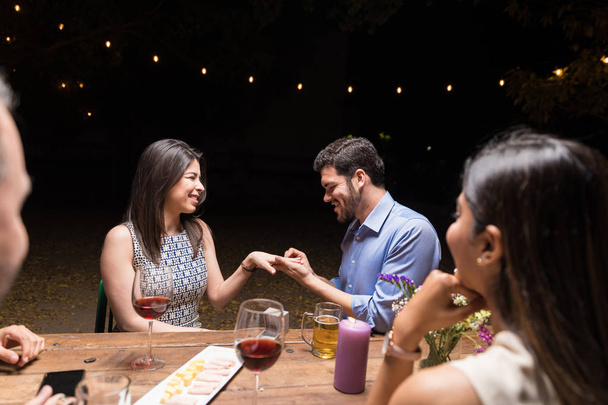 Νεαρός άνδρας βάζει δαχτυλίδι στο δάχτυλο της φίλης του σε δείπνο με φίλους στο εστιατόριο - Φωτογραφία, εικόνα