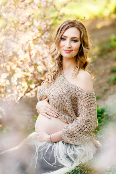 Beautiful pregnant woman sitting near the cherry blossom. Pregnant happy pretty woman in blossom white garden. - Foto, Bild