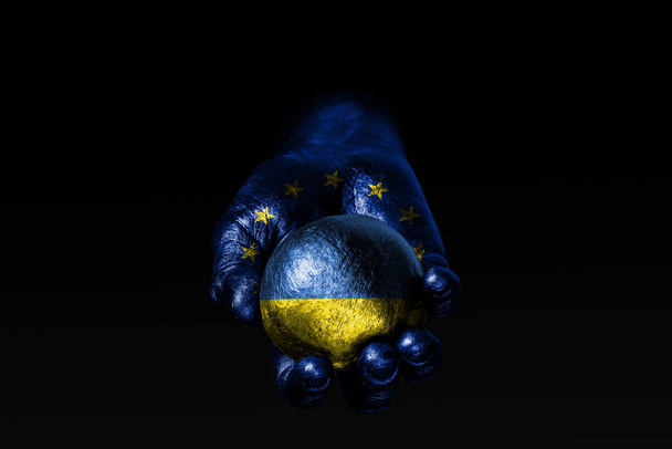 Ręka z wyciągnięte flagi UE posiada piłkę z wyciągnięte flagi Ukrainy, znak wpływów, presji lub ochrony i ochrona. - Zdjęcie, obraz