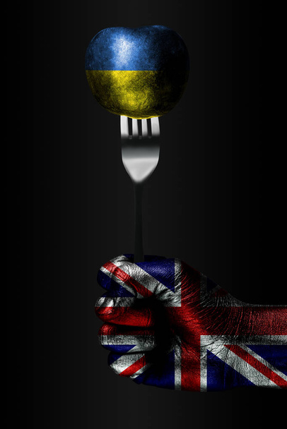 A hand with a drawn Great Britain flag holds a fork, on which is a ball with a drawn Ukraine flag, a sign of influence, pressure, grip and anecxia. - Photo, Image