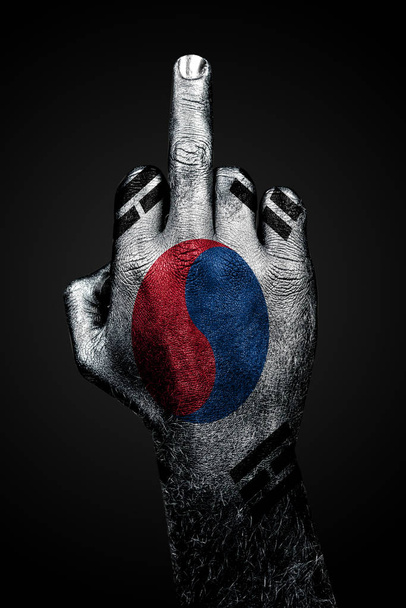 Een hand met een geschilderde vlag van Zuid-Korea toont de middelvinger, een teken van agressie, tegen een donkere achtergrond - Foto, afbeelding