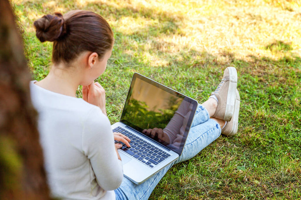 Jonge vrouw zittend op groen gras gazon in stadspark op laptop pc computer werkt. Freelance bedrijfsconcept - Foto, afbeelding