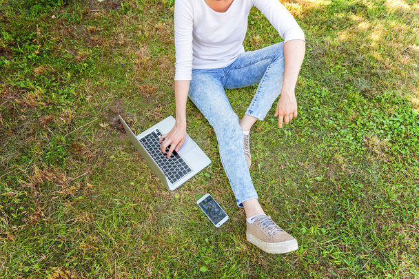 Woman legs on green grass lawn in city park, hands working on laptop pc computer. Freelance business concept - Zdjęcie, obraz
