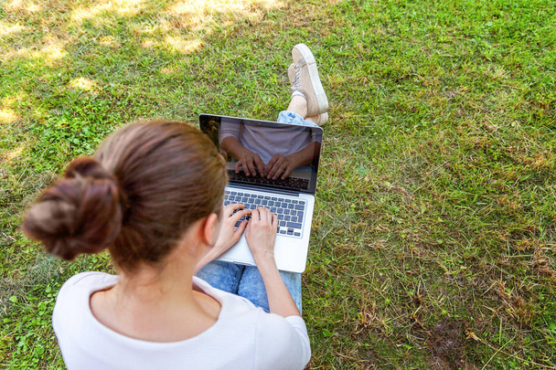 Młoda kobieta siedzi na zielony trawa trawnik w parku miejskim, w pracy na komputerze pc laptop. Koncepcja działalności freelancerskiej - Zdjęcie, obraz