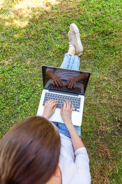 Nuori nainen istuu vihreä ruoho nurmikko kaupungin puistossa työskentelee kannettavan tietokoneen. Freelance-liiketoiminnan käsite
 - Valokuva, kuva