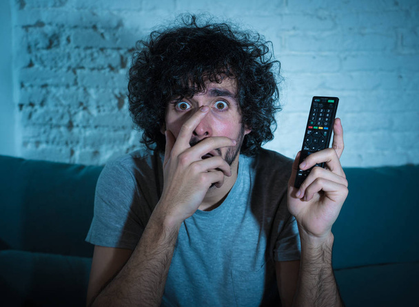 Lifestyle portrait of young man feeling scared and shocked making fear, anxiety gestures while watching television holding remote control. In horror and violence on TV and internet and mass media. - Photo, Image