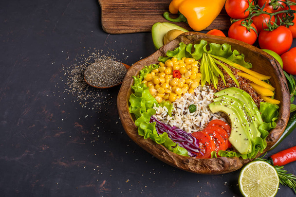 Wooden bowl with chickpea, avocado, wild rice, quinoa, bell pepper, tomatoes, greens, cabbage, lettuce on dark stone table and wooden spoon with chia seeds with copy space. - 写真・画像