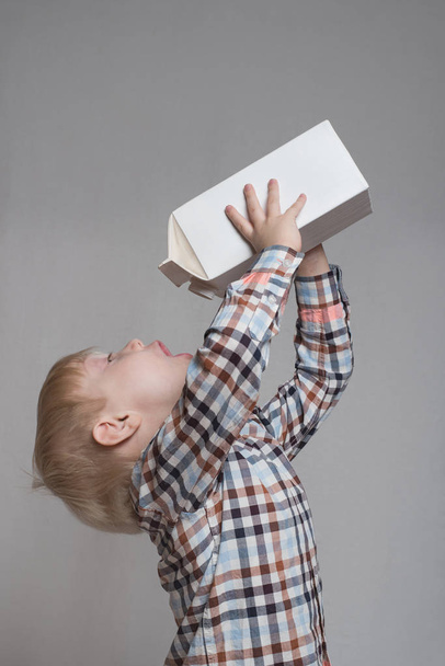 Little blond boy drinks from a large white package. Light background - Photo, Image