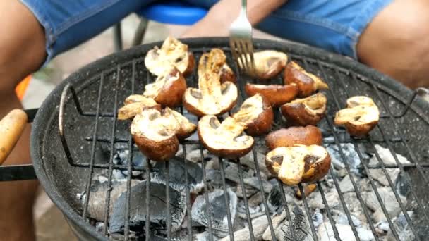 Grilled food. Man turn over large delicious champignon mushrooms, which roast over the coals on barbecue, on a small outdoor grill on a metal grate. Weekend relaxation concept. - Footage, Video