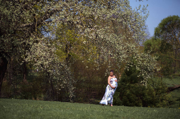 Young traveler pregnant woman walking, running, turning around and enjoys her leisure free time in a park with blossoming sakura cherry trees wearing a summer light long dress with flower pattern - Photo, Image
