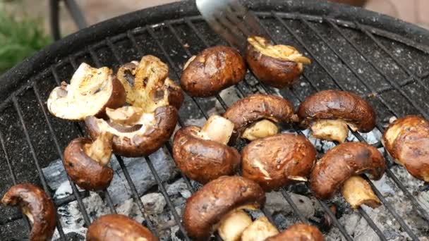 Grilled food. Man turn over large delicious champignon mushrooms, which roast over the coals on barbecue, on a small outdoor grill on a metal grate. Weekend relaxation concept. - Footage, Video