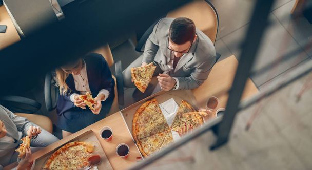 Bovenaanzicht van zakenmensen in formele slijtage zittend aan tafel en pizza voor de lunch. - Foto, afbeelding