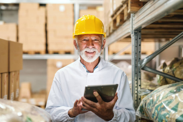 Trabajador senior sonriente en uniforme y con casco amarillo en la cabeza sosteniendo la tableta y mirando a la cámara mientras está de pie en el almacenamiento
. - Foto, imagen