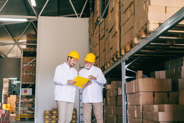 Two workers dressed in uniforms and with helmets on heads looking at documents and discussing while standing in storage. - Photo, Image