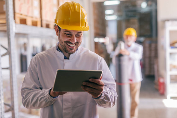 Close up of young Caucasian smiling worker with helmet on head using tablet for work while standing in warehouse. - Photo, Image
