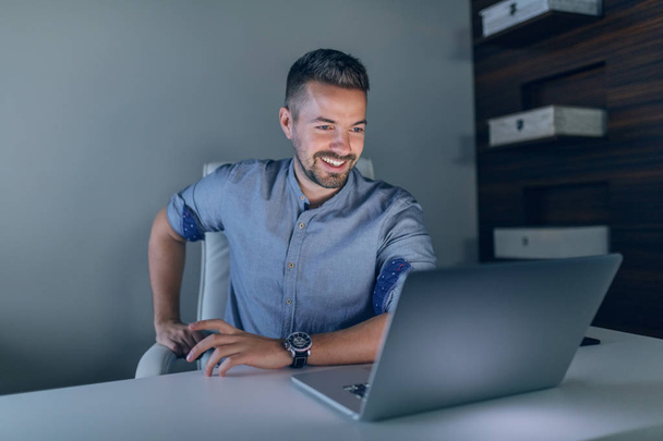Χαμογελαστός εργαζόμενος Καυκάσιος επιχειρηματίας κοιτάζοντας το laptop, ενώ κάθονται στο γραφείο αργά τη νύχτα. - Φωτογραφία, εικόνα