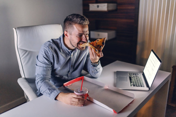 Joven arquitecto caucásico comiendo pizza y bebiendo jugo en la oficina. En el portátil de escritorio. Concepto de trabajo nocturno
. - Foto, Imagen