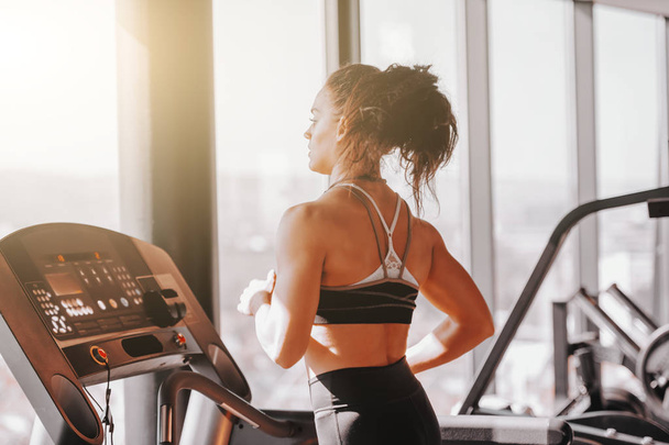 Young sporty muscular brunette with ponytail running on treadmill. Backs turned. Train like an athlete, eat like nutritionist, sleep like a baby and win like a champion. - Photo, Image