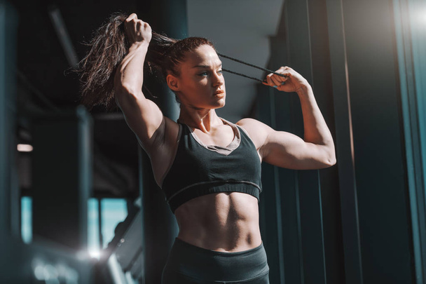 Young Caucasian strong muscular female bodybuilder tying hair while standing in gym next to window. If you're tired of starting over, stop giving up. - Photo, Image