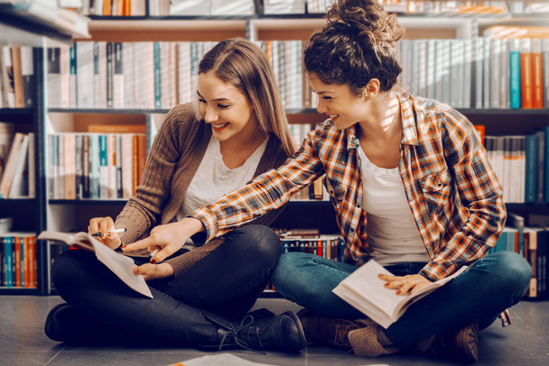 Két kaukázusi csinos női diák ül, lábait keresztbe a könyvtár és a tanulás a vizsgákra. Lány göndör hajú mutatott a válasz a könyvben. - Fotó, kép