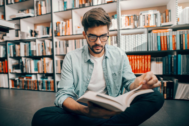 Mixed race male student sitting on library floor with legs crossed and reading book. In background books on shelves. - Photo, Image