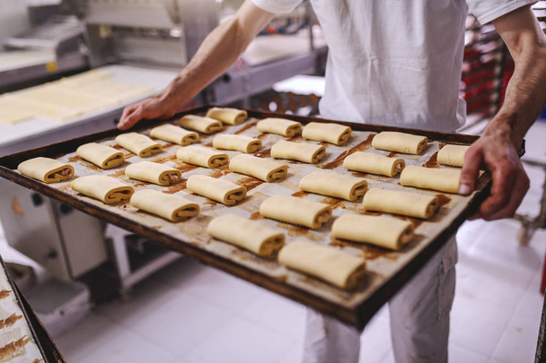 panadero en uniforme blanco celebración de hornear orar con crudo dulce deliciosa pastelería mientras está de pie en panadería
. - Foto, imagen