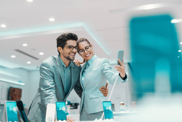 Νεαρό ελκυστικό πολυπολιτισμικό ζευγάρι ντυμένο κομψό λαμβάνοντας selfie με νέο έξυπνο τηλέφωνο στο κατάστημα τεχνολογίας. Γυναίκα κρατά έξυπνο τηλέφωνο ενώ ο άντρας την αγκαλιάζει. - Φωτογραφία, εικόνα