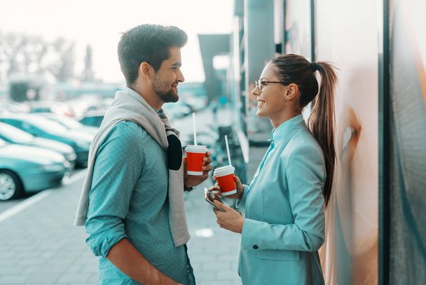 Усміхнена мультикультурна пара одягнена елегантно стоячи на відкритому повітрі, дивлячись один на одного і п'ючи каву, щоб піти. Жінка тримає планшет
. - Фото, зображення
