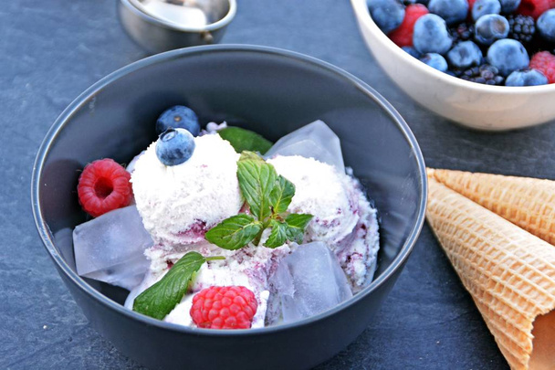 A dark bowl full of wild berry ice cream balls with mint leaves and ice cream waffles next to it on a dark kitchen surface - concept with refreshing fruit ice for the summer - Photo, Image