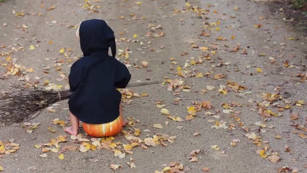child in black sorcerer or witch suit with broom sits on pumpkin and looks on falling autumn leaves, copy space - Footage, Video