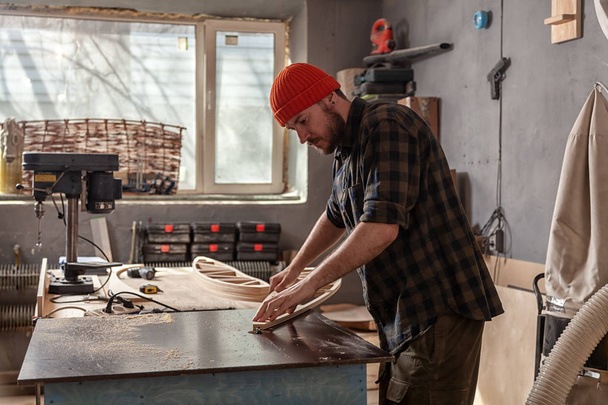 Egy fiatalember Ács építő munkaruhák feldolgozása egy fából készült deszka a marógép a műhelyben, körül sok felszerelés, fa deszkák. Lakásjavítási koncepciók. - Fotó, kép