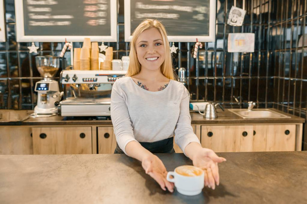 Young smiling barista girl with cup of freshly prepared coffee. Blond woman in an apron, near the bar counter in coffee shop - Valokuva, kuva