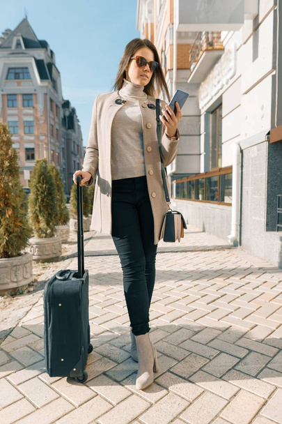 Portrait of traveling young woman with mobile phone and suitcase, fashionable girl on the city street, wearing warm coat, sunny autumn day - Photo, Image