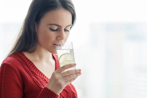 Young beautiful woman in red sweater with glass of water with lime, woman stands near window in skyscraper on cloudy day. Healthy drink, natural antioxidant, vitamins in urban life - Photo, image
