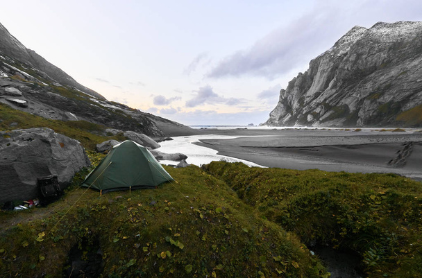 Green tent on a hill at Bunes Beach on Lofoten Islands in Norway. The tent is standing on a grass hill next to a big rock. In the background there are mountains, the beach and the setting sun. - Photo, Image
